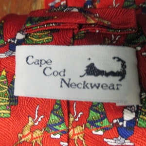 May include: Red Cape Cod Neckwear tie with a festive Christmas pattern. The tie features illustrations of Santa Claus skiing, decorated Christmas trees, and reindeer. The label reads "Cape Cod Neckwear".