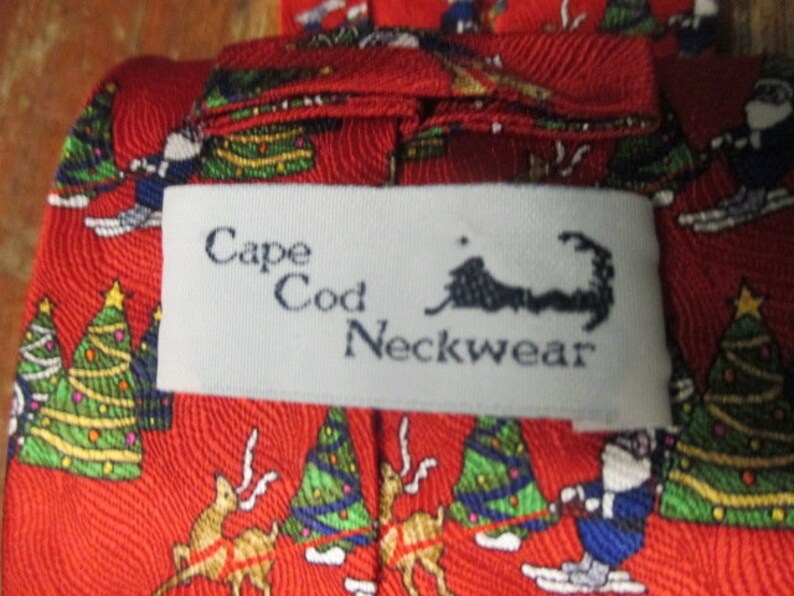 May include: Red Cape Cod Neckwear tie with a festive Christmas pattern. The tie features illustrations of Santa Claus skiing, decorated Christmas trees, and reindeer. The label reads "Cape Cod Neckwear".