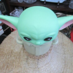 May include: A green figure with large ears and black eyes, reminiscent of a popular character. The figure has a light green head and ears with pink inner ears. It is wearing a beige outfit and is sitting on a light brown surface.