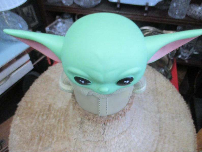 May include: A green figure with large ears and black eyes, reminiscent of a popular character. The figure has a light green head and ears with pink inner ears. It is wearing a beige outfit and is sitting on a light brown surface.