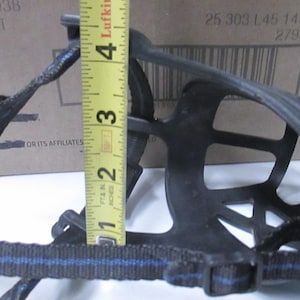 May include: Black dog muzzle with a cage-like design. The muzzle has a strap with blue stripes and a buckle. A yellow tape measure shows the muzzle is approximately 3 inches tall.