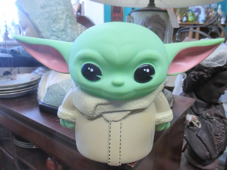 May include: A green figurine of a character with large black eyes, large ears with pink insides, and a tan coat. The figurine is a stylized representation of a popular character from a science fiction franchise.