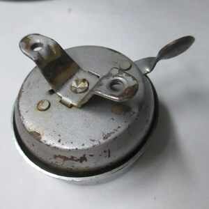 May include: A vintage circular metal bicycle bell with a weathered silver finish. The bell features a central mounting bracket with two screw holes and a small lever arm extending from the side. The surface shows signs of oxidation and wear.
