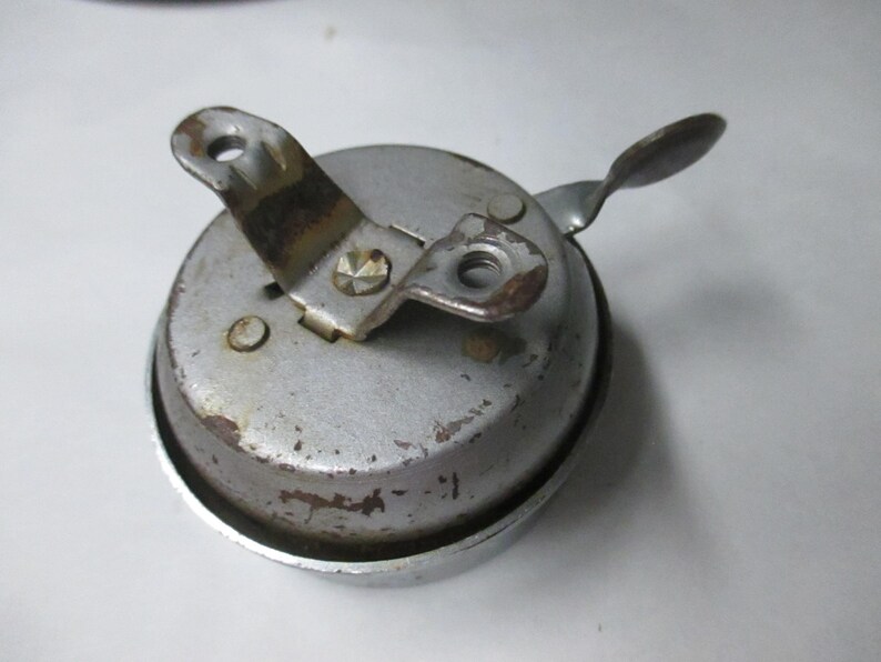 May include: A vintage circular metal bicycle bell with a weathered silver finish. The bell features a central mounting bracket with two screw holes and a small lever arm extending from the side. The surface shows signs of oxidation and wear.