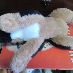 May include: A plush dog toy in tan, black, and white, lying on a red book. The toy has floppy ears and a soft, furry texture. The book has the word "Follow" and "ND" visible.