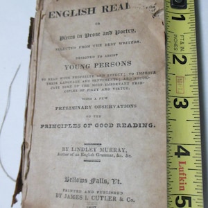 May include: An antique book titled "The English Real" with text in black ink on aged paper. The book is measured with a yellow tape measure, showing it is approximately 5 inches tall. The book's cover includes the year 1827.