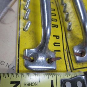 May include: A close-up of a silver-colored door pull with a curved handle, mounted on a yellow card. The card has the words "DOOR PULL" printed on it. A ruler shows the door pull is approximately 1 inch in length.