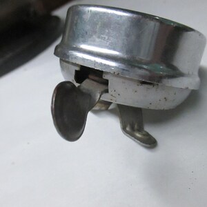 May include: A vintage chrome-plated bicycle bell with a rounded top and a side-mounted metal thumb lever. The bell features a metallic silver finish with visible signs of wear and oxidation on the lower casing and the activation mechanism.