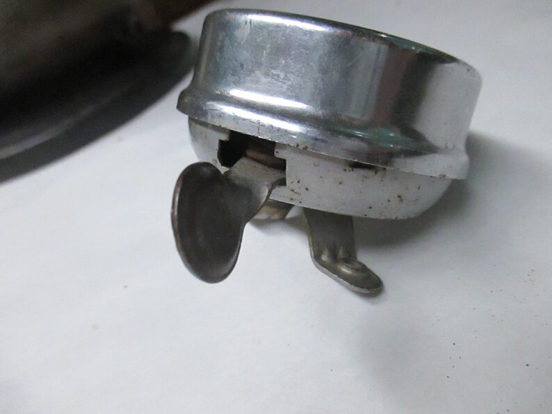 May include: A vintage chrome-plated bicycle bell with a rounded top and a side-mounted metal thumb lever. The bell features a metallic silver finish with visible signs of wear and oxidation on the lower casing and the activation mechanism.