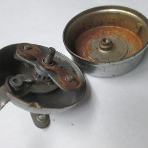 May include: A disassembled chrome bicycle bell consisting of two main parts. The base features an internal gear mechanism with a lever and visible rust. The separate circular top cover shows oxidation and rust on its interior surface.