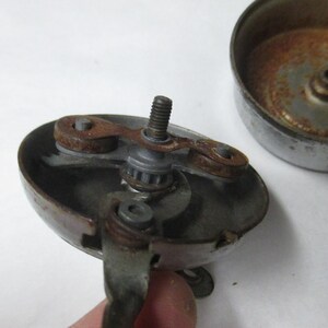 May include: A disassembled metal bicycle bell featuring a circular base with a central threaded screw, a rotating internal mechanism with two small brown rusted components, and a separate detached metallic dome cover showing signs of oxidation.