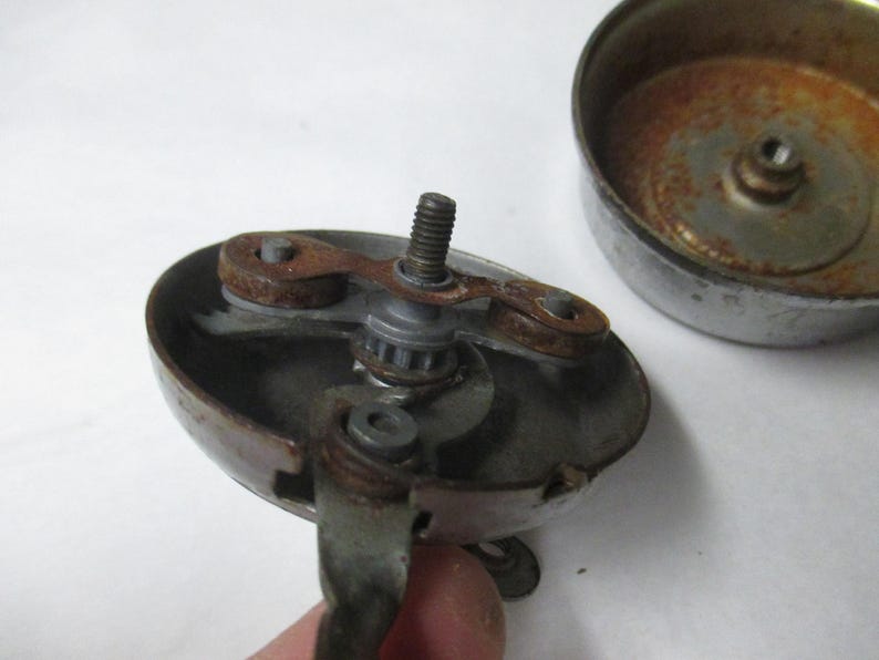 May include: A disassembled metal bicycle bell featuring a circular base with a central threaded screw, a rotating internal mechanism with two small brown rusted components, and a separate detached metallic dome cover showing signs of oxidation.