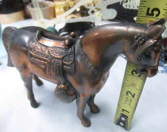 May include: A copper-toned metal horse figurine with an ornate saddle and bridle. A yellow measuring tape is positioned against the horse's neck, indicating a height of approximately 5 inches from the base to the top of the head.