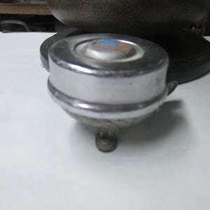 May include: A round, chrome-plated bicycle bell with a smooth, reflective metallic finish. The top center features a small, circular inset with a blue and brown rectangular graphic. A small metal lever extends from the side for activation.