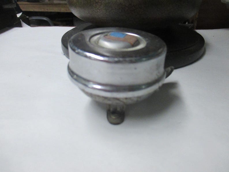 May include: A round, chrome-plated bicycle bell with a smooth, reflective metallic finish. The top center features a small, circular inset with a blue and brown rectangular graphic. A small metal lever extends from the side for activation.