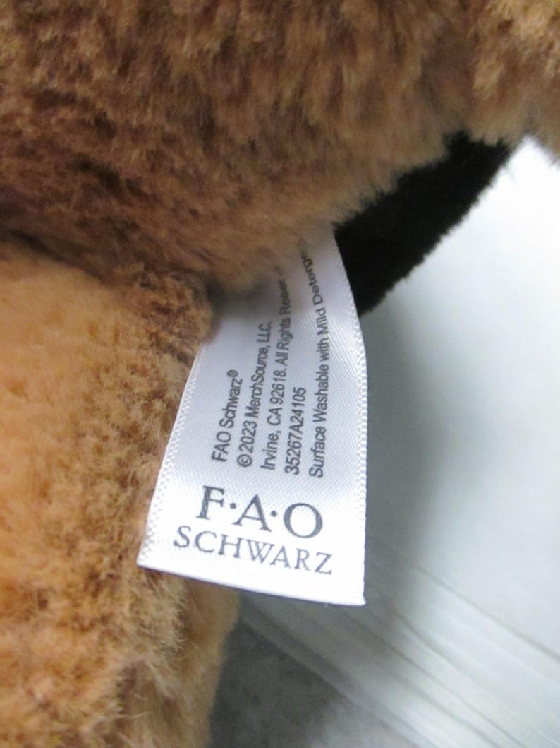 May include: Close-up of a plush toy with brown fur and a black patch. A white tag is attached, displaying the "FAO Schwarz" logo and washing instructions.