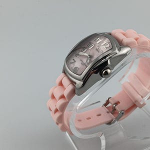 Puede incluir: Un reloj plateado con esfera rosa y correa de silicona rosa a juego. El reloj tiene una esfera rectangular con agujas y n&uacute;meros plateados. La correa se sujeta con una hebilla plateada. El reloj se exhibe sobre un soporte transparente.