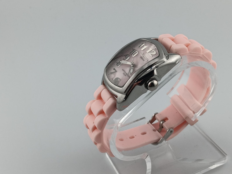 Puede incluir: Un reloj plateado con esfera rosa y correa de silicona rosa a juego. El reloj tiene una esfera rectangular con agujas y n&uacute;meros plateados. La correa se sujeta con una hebilla plateada. El reloj se exhibe sobre un soporte transparente.