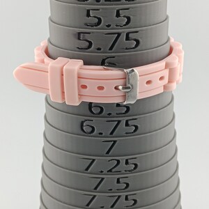 Puede incluir: Una correa de reloj rosa claro est&aacute; sujeta a un medidor de anillos gris. El medidor de anillos tiene medidas de 5,25 a 8,25. La correa del reloj tiene una hebilla plateada.