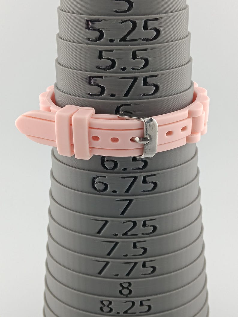 Puede incluir: Una correa de reloj rosa claro est&aacute; sujeta a un medidor de anillos gris. El medidor de anillos tiene medidas de 5,25 a 8,25. La correa del reloj tiene una hebilla plateada.