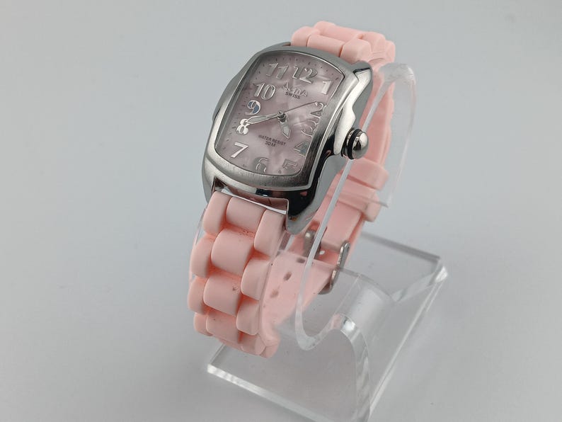 Puede incluir: Un reloj de pulsera rosa con caja plateada y correa de silicona rosa. La esfera del reloj es rosa claro con n&uacute;meros y agujas plateadas. El reloj se exhibe en un soporte de pl&aacute;stico transparente.
