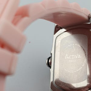 Puede incluir: Primer plano de un reloj plateado con una correa rosa claro. La esfera del reloj no es visible. La parte trasera del reloj tiene la marca "Activa Swiss" impresa. La correa est&aacute; hecha de un material suave y flexible.