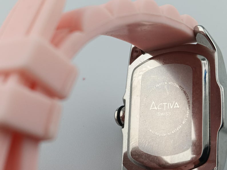 Puede incluir: Primer plano de un reloj plateado con una correa rosa claro. La esfera del reloj no es visible. La parte trasera del reloj tiene la marca "Activa Swiss" impresa. La correa est&aacute; hecha de un material suave y flexible.