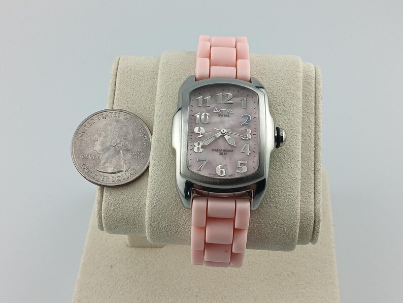 Puede incluir: Un reloj Activa Swiss plateado con una correa y esfera rosa claro. El reloj tiene una esfera rectangular con n&uacute;meros y agujas plateadas. Una moneda est&aacute; al lado del reloj para comparar el tama&ntilde;o.