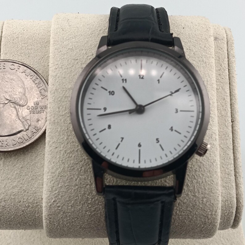 May include: A black wristwatch with a white face and black hands. The watch has a black leather band and a silver quarter is next to the watch. The watch face has black numbers and markers.