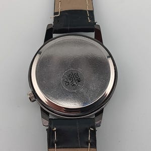 May include: Close-up of a wristwatch with a black leather band. The watch has a silver-colored case and a round, metallic back. The back of the watch has the text "QC PASS" and the number "1602" printed on it.