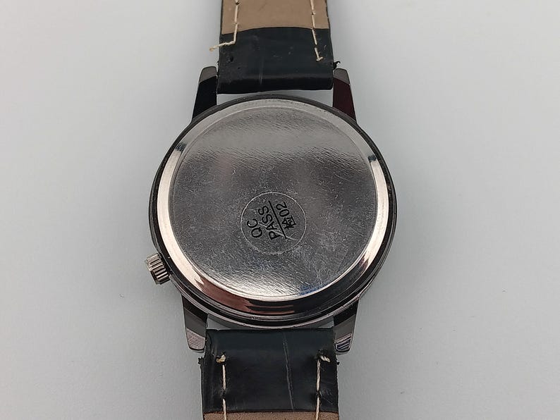 May include: Close-up of a wristwatch with a black leather band. The watch has a silver-colored case and a round, metallic back. The back of the watch has the text "QC PASS" and the number "1602" printed on it.