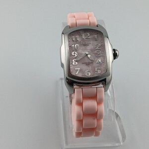 Puede incluir: Un reloj Activa Swiss plateado con esfera rectangular y correa rosa claro. La esfera tiene un acabado nacarado con n&uacute;meros y agujas plateadas. La correa est&aacute; hecha de eslabones rosa claro.
