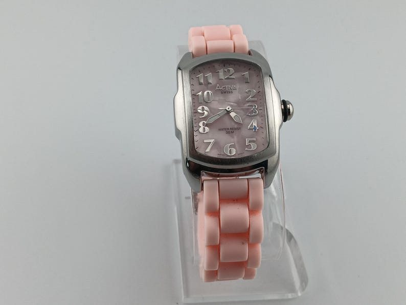 Puede incluir: Un reloj Activa Swiss plateado con esfera rectangular y correa rosa claro. La esfera tiene un acabado nacarado con n&uacute;meros y agujas plateadas. La correa est&aacute; hecha de eslabones rosa claro.