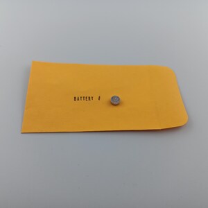 May include: A small, silver button cell battery rests on a yellow envelope. The envelope is labeled "BATTERY #" in black text. The background is a plain, light gray surface.