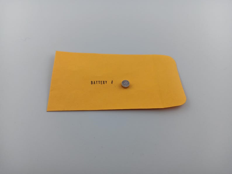May include: A small, silver button cell battery rests on a yellow envelope. The envelope is labeled "BATTERY #" in black text. The background is a plain, light gray surface.