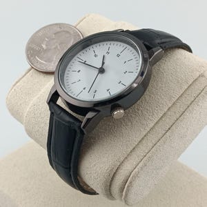 May include: A black wristwatch with a white face and black leather band. The watch has a round face with black numerals and hands. A quarter is next to the watch. The watch is displayed on a beige surface.