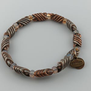 Può includere: Un braccialetto elasticizzato con perline di metallo attorcigliate color rame e argento alternate, intervallate da piccole perline iridescenti. È attaccato un piccolo ciondolo rotondo in rame con la scritta "VINTAGE". Il braccialetto è progettato per una vestibilità comoda.