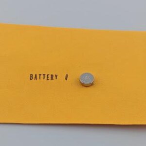 Puede incluir: Una peque&ntilde;a pila de bot&oacute;n plateada descansa sobre una superficie rectangular de color amarillo brillante. La palabra "BATTERY" est&aacute; impresa en negro, junto a un peque&ntilde;o s&iacute;mbolo. El fondo es gris claro.