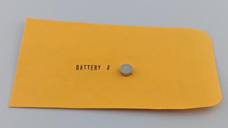 Puede incluir: Una peque&ntilde;a pila de bot&oacute;n plateada descansa sobre una superficie rectangular de color amarillo brillante. La palabra "BATTERY" est&aacute; impresa en negro, junto a un peque&ntilde;o s&iacute;mbolo. El fondo es gris claro.