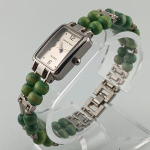 Puede incluir: Reloj de cuarzo plateado con esfera rectangular, esfera blanca y el texto "Cote d'Azur". El reloj tiene una pulsera de cuentas verdes y una correa de metal plateado. El reloj se exhibe en un soporte transparente.