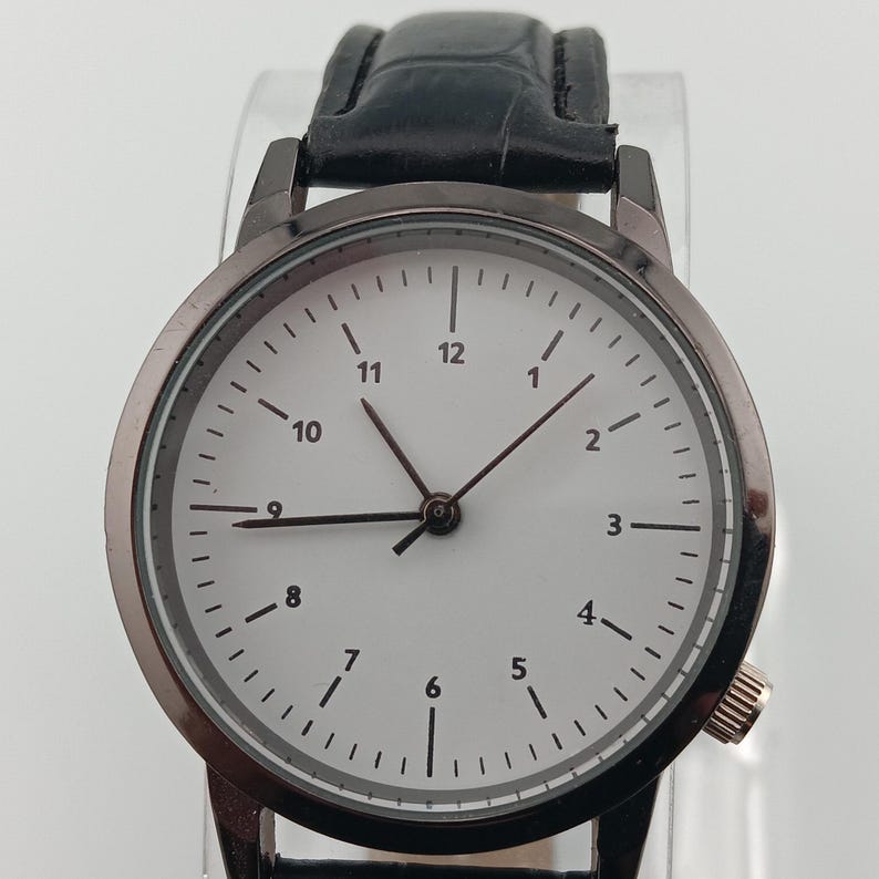 May include: A round wristwatch with a white face and black numerals. The watch has black hands and a black leather band. The watch case is a dark gray color. The watch has a small gold-colored crown on the side.