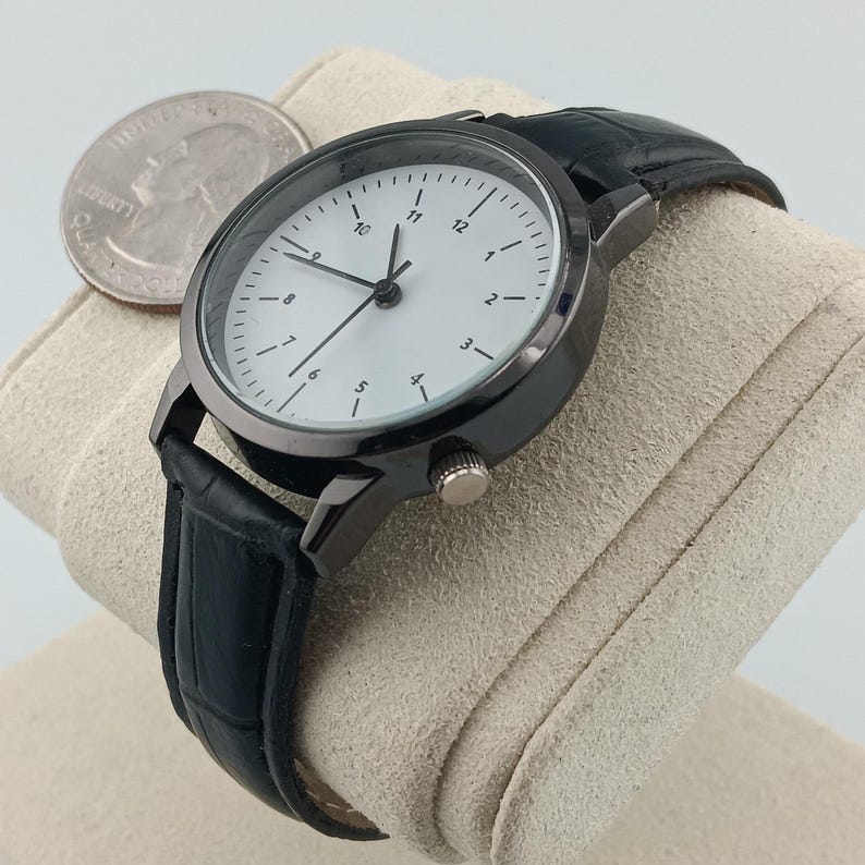May include: A black wristwatch with a white watch face and black leather band. The watch has a dark gray metal casing and a small dial on the side. A quarter is next to the watch.