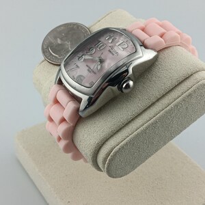 Puede incluir: Un reloj plateado con una correa rosa y una esfera rosa. El reloj tiene una esfera rectangular con n&uacute;meros y agujas blancas. La correa est&aacute; hecha de un material suave y flexible. Una moneda est&aacute; colocada junto al reloj.