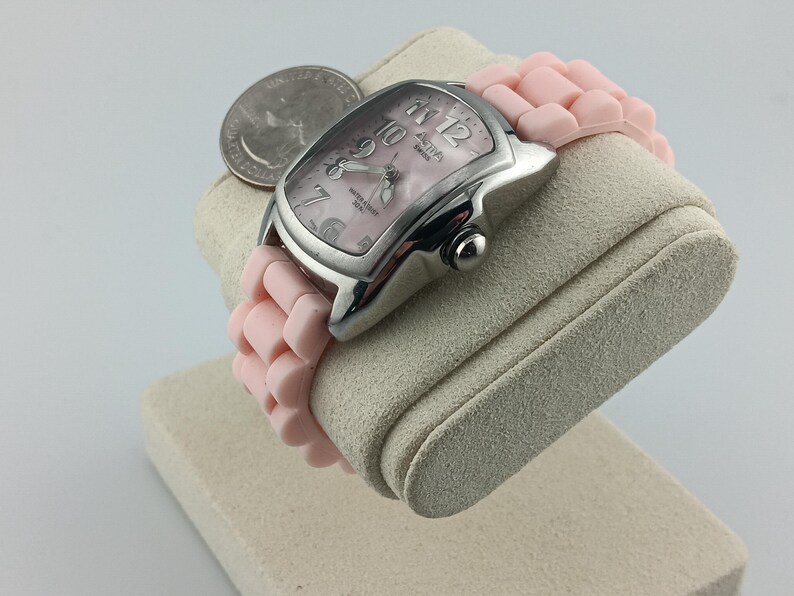 Puede incluir: Un reloj plateado con una correa rosa y una esfera rosa. El reloj tiene una esfera rectangular con n&uacute;meros y agujas blancas. La correa est&aacute; hecha de un material suave y flexible. Una moneda est&aacute; colocada junto al reloj.