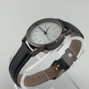 May include: A black wristwatch with a round, dark gray case and a white face featuring black numerals and hands. The watch has a black leather band with a buckle closure. The watch is displayed on a clear, curved surface.