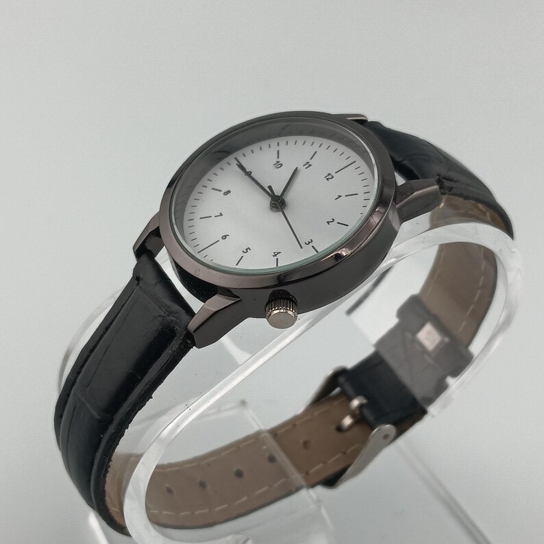 May include: A black wristwatch with a round, dark gray case and a white face featuring black numerals and hands. The watch has a black leather band with a buckle closure. The watch is displayed on a clear, curved surface.