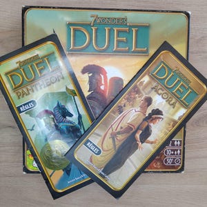 Peut inclure: Un jeu de société "7 Wonders Duel" avec des extensions. La boîte principale est vert clair avec des lettres dorées. Deux extensions, "Pantheon" et "Agora", sont visibles, chacune avec des illustrations uniques et le mot "Règles".