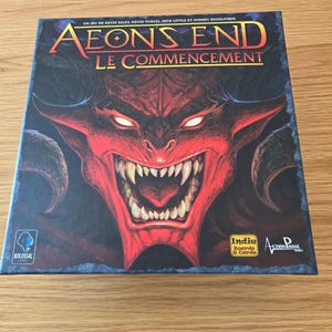 Peut inclure: Une boîte de jeu de société carrée intitulée Aeon's End Le Commencement illustrée d'un grand démon rouge à cornes avec des yeux brillants et des dents pointues. Le haut indique les noms des concepteurs et le bas affiche les logos de Kolossal Games, Indie Boards & Cards et Action Phase Games.