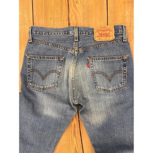 Könnte beinhalten: Verblasste blaue Jeans mit dem Logo-Patch von Levi Strauss & Co. am hinteren Bund. Die Jeans haben ein klassisches Fünf-Taschen-Design mit der charakteristischen Levi's-Bogennaht auf den Gesäßtaschen. Ein kleines rotes Etikett ist sichtbar.