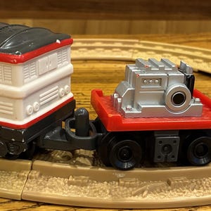 May include: A toy train set with a red, white, and black engine, a silver cargo car, and a red caboose carrying a small figure of a scientist. The train is on a tan circular track.